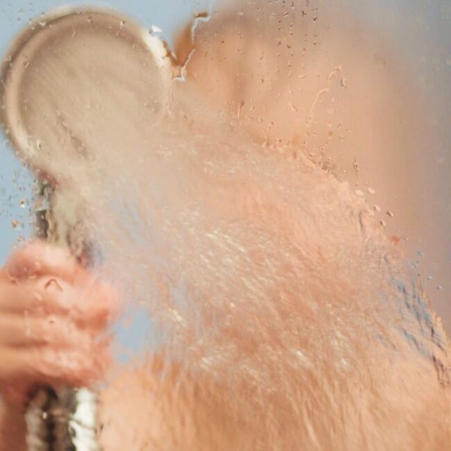 Personne utilisant un pommeau de douche sur une vitre remplie de buée.